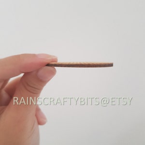 May include: A thin, round wooden craft piece with a light brown surface and a darker brown edge. The text "RAINSCRAFTYBITS@ETSY" is printed in a light green color. The piece is held between fingers against a white background.
