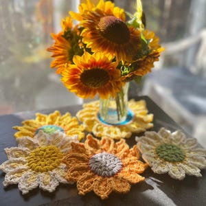 May include: Four crocheted sunflower coasters in yellow, white, and green. The coasters are arranged on a dark surface with a vase of sunflowers in the background.