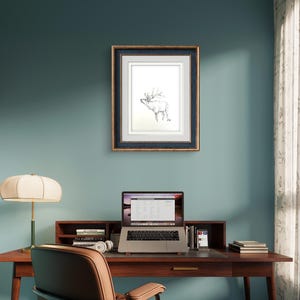 May include: A framed black and white line drawing of an elk with antlers, hanging on a teal wall above a wooden desk with a laptop, books, and a lamp.