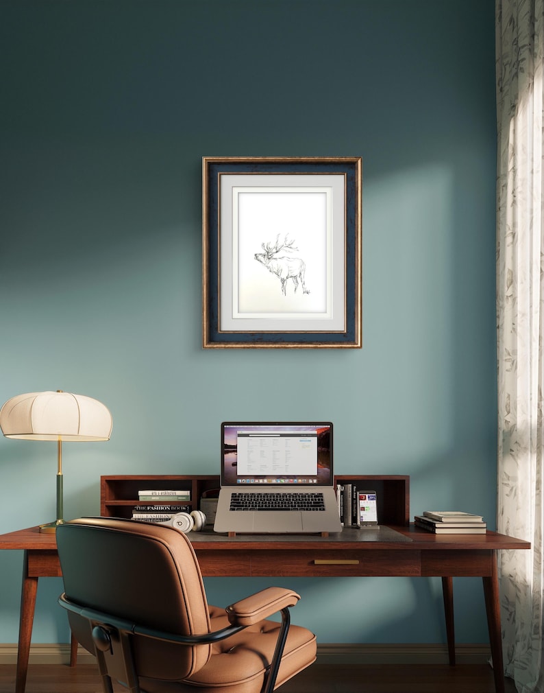 May include: A framed black and white line drawing of an elk with antlers, hanging on a teal wall above a wooden desk with a laptop, books, and a lamp.