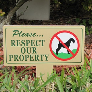 May include: A tan sign with green text and a red circle with a black silhouette of a dog with a red line through it. The sign reads "Please... RESPECT OUR PROPERTY".