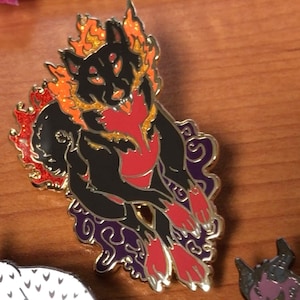 May include: A black wolf enamel pin with fiery orange and red accents. The wolf is depicted with flames around its head and body, set against a purple and gold decorative border. The pin has a gold metallic outline.