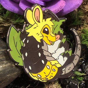 May include: A black and yellow enamel pin featuring a cartoon animal with a long snout, large ears, and a bushy tail. The animal is surrounded by green leaves and is set against a background of moss, rocks, and flowers.