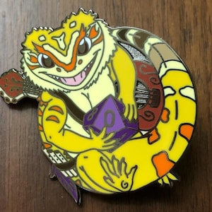 May include: An enamel pin featuring a cartoon lizard playing a lute. The lizard is yellow and orange with a purple and gold sash and a purple die in its hand.