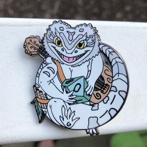 May include: White enamel pin of a bearded dragon holding a green d20 die and a lute. The dragon is wearing a brown strap across its chest and has a white and grey tail.