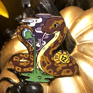 May include: A gold and brown enamel pin featuring a snake with a green and purple background. The snake is coiled around a yellow and black dice with the number 12 on it. The snake has a green and purple slime dripping from its mouth.
