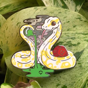 May include: A yellow and white snake enamel pin with a green plant and a red and white dice. The snake is curled around the dice and the plant is growing out of its mouth.