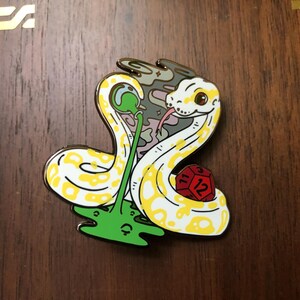 May include: A yellow and white snake with a red and yellow dice in its coils. The snake is curled around a green plant with a purple and grey background. The snake has a black eye and a pink tongue.