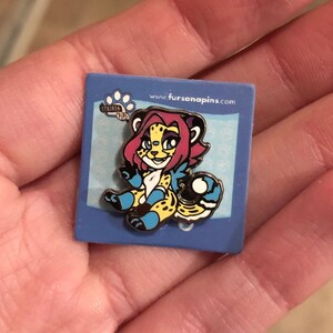 Puede incluir: Un pin de esmalte azul y amarillo con un guepardo de dibujos animados con pelo rosa y ojos azules. El guepardo est&aacute; sosteniendo una pelota azul y blanca. El pin est&aacute; sobre un fondo azul con el texto "www.fursanapins.com" y una huella de pata.