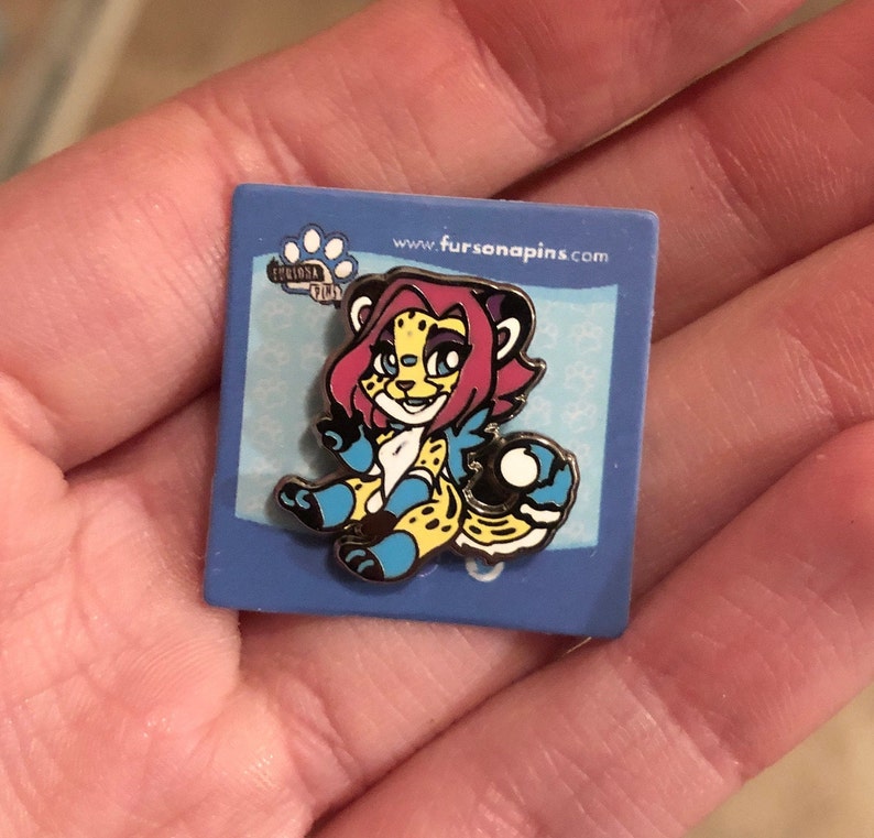 Puede incluir: Un pin de esmalte azul y amarillo con un guepardo de dibujos animados con pelo rosa y ojos azules. El guepardo est&aacute; sosteniendo una pelota azul y blanca. El pin est&aacute; sobre un fondo azul con el texto "www.fursanapins.com" y una huella de pata.