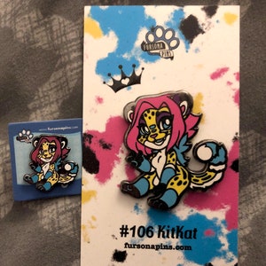 Puede incluir: Un pin de esmalte colorido con un guepardo de dibujos animados con pelo rosa, ojos azules y un pelaje manchado de amarillo y negro. El pin est&aacute; en una tarjeta blanca con una corona negra y el texto "#106 Kitkat fursonapins.com".