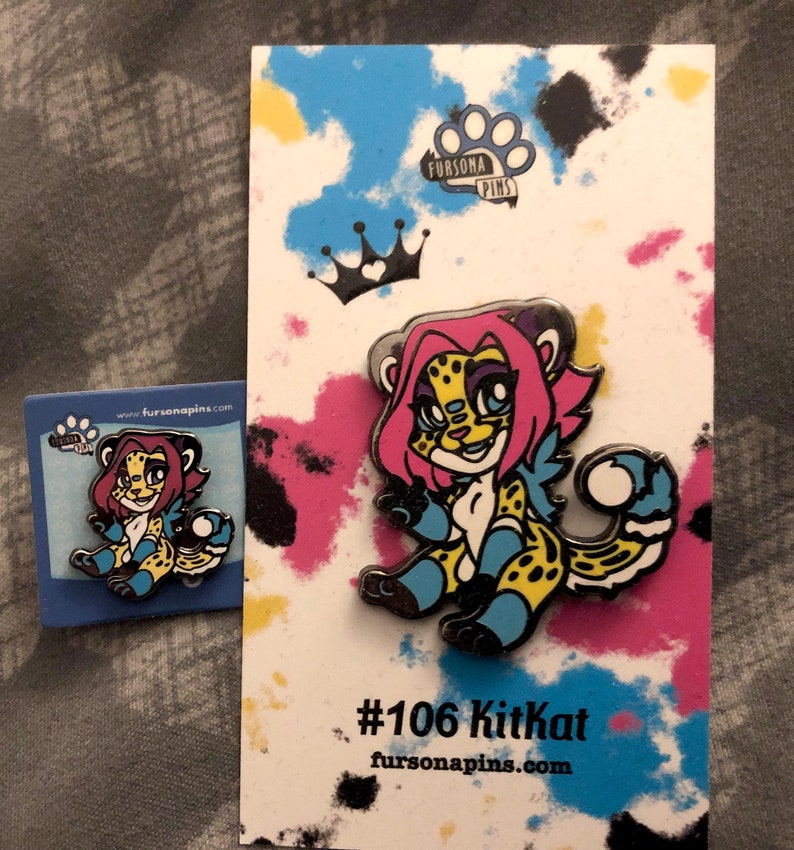 Puede incluir: Un pin de esmalte colorido con un guepardo de dibujos animados con pelo rosa, ojos azules y un pelaje manchado de amarillo y negro. El pin est&aacute; en una tarjeta blanca con una corona negra y el texto "#106 Kitkat fursonapins.com".