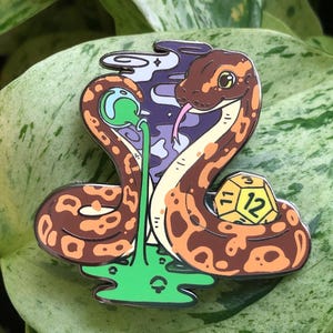 May include: A brown and orange snake with a green and purple background. The snake is holding a yellow 12-sided die. The snake is wearing a green and purple hat.