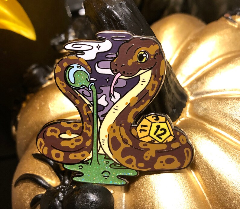May include: A gold and brown enamel pin featuring a snake with a green and purple background. The snake is coiled around a yellow and black dice with the number 12 on it. The snake has a green and purple slime dripping from its mouth.