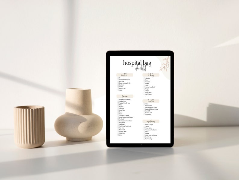 May include: A digital tablet displaying a hospital bag checklist with sections for "Mom", "Baby", "For the Trip", and "Essentials". The checklist includes items such as toiletries, clothing, and documents. The tablet is on a white surface with two beige vases.