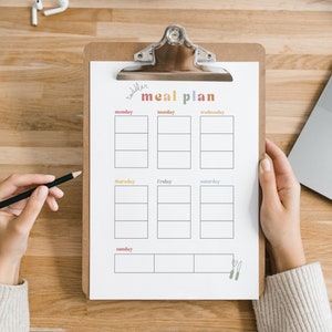 May include: A weekly meal planner on a clipboard with a brown wooden background. The planner has a title that says "weekly meal plan" and has spaces for each day of the week.