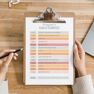May include: A clipboard with a printed daily schedule titled "Grayson's Daily Schedule". The schedule includes times and activities like "Free Play", "Lunch", and "Bedtime Routine". A hand holds a pencil, and a laptop and earbuds are in the background.