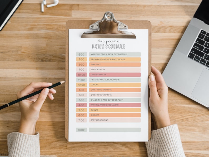 May include: A clipboard with a printed daily schedule titled "Grayson's Daily Schedule". The schedule includes times and activities like "Free Play", "Lunch", and "Bedtime Routine". A hand holds a pencil, and a laptop and earbuds are in the background.