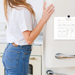 May include: A white magnetic weekly dinner menu, titled "Baby Family Weekly Dinner Menu", is attached to a white refrigerator. The menu has sections for each day of the week, with space to write meal ideas. A black magnet holds the menu in place.