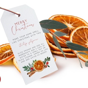 Op de afbeelding: Een wit cadeau-etiket met rode tekst die "Vrolijk Kerstfeest" luidt. Het etiket bevat instructies om je huis naar Kerstmis te laten ruiken met een "Stoofpotpourri" recept. Het etiket is versierd met een handgetekende illustratie van sinaasappels, kaneelstokjes en groen.