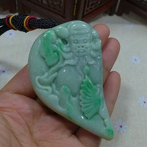 May include: A green jade pendant carved with a detailed depiction of a mythical figure. The pendant is shaped like a heart and has a smooth, polished surface.