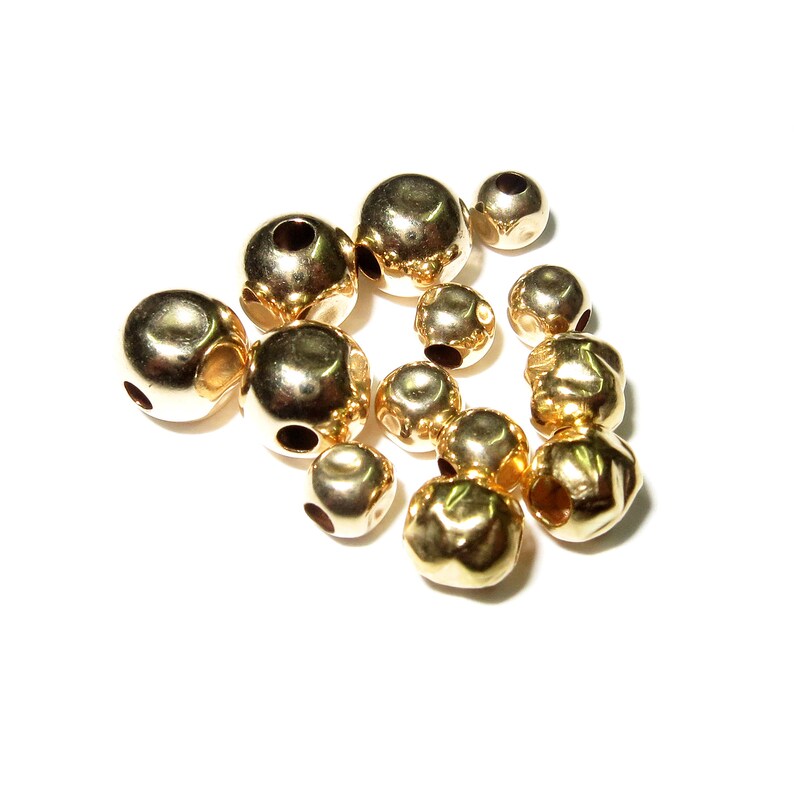 May include: A group of gold-colored metal beads with a hole in the center of each bead. The beads are arranged in a loose pile on a white surface.