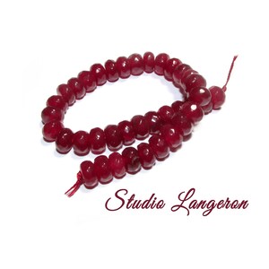 May include: A strand of faceted, deep red, circular beads arranged in a loose oval shape on a white background. The beads have a translucent quality and a polished finish. The text Studio Langeron is printed in a dark red script font below the beads.
