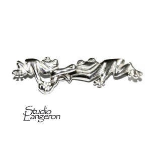 Puede incluir: Un broche de plata con dos ranas entrelazadas. Las ranas están mirando en direcciones opuestas y sus patas están entrelazadas. El broche está hecho de plata y tiene un acabado pulido.