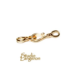 May include: A gold-colored jewelry clasp with a hook and eye closure. The clasp is made of a shiny metal and is designed to connect two pieces of jewelry. The text "Studio L'Angeron" is printed in gold.