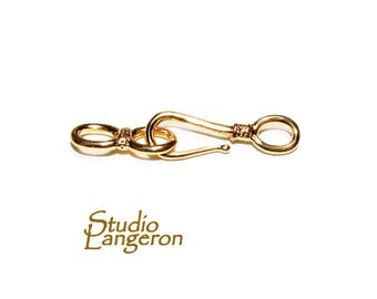 May include: A gold-colored jewelry clasp with a hook and eye design. The clasp has a curved hook and two circular loops, with intricate detailing. The Studio Langeron logo is visible in the bottom left corner.