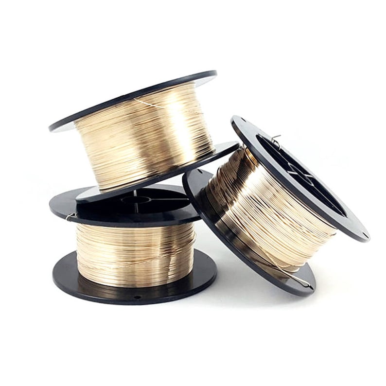 May include: Three spools of gold-colored wire are stacked and leaning against each other. The wire is tightly wound on black plastic spools. The image is well-lit, highlighting the metallic sheen of the wire, suitable for jewelry making or crafting.