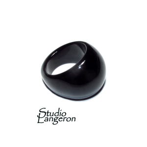Puede incluir: Un anillo negro brillante con forma redondeada y abovedada. El anillo es de color liso y tiene una superficie lisa y reflectante. Es un accesorio de moda.