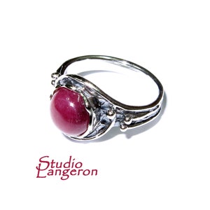 Puede incluir: Un anillo de plata con una piedra preciosa roja oscura engastada en una banda texturizada y detallada. El anillo tiene un diseño delicado e intrincado con pequeñas cuentas de plata.
