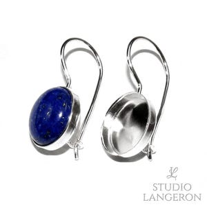 Op de afbeelding: Een paar zilverkleurige oorbellen met lange, gebogen oorhaken. De ene oorbel bevat een gladde, ovaalvormige diepblauwe lapis lazuli steen in een metalen zetting, terwijl de andere een lege zilverkleurige metalen zetting van dezelfde vorm is.