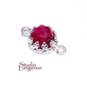 Puede incluir: Un conector de plata con una piedra preciosa roja en un engaste de corona. El conector tiene dos bucles para sujetarlo a joyas.