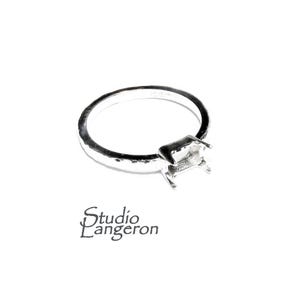 Peut inclure: Une bague en argent avec un sertissage carré, prête à recevoir une pierre précieuse. L'anneau a un aspect texturé et martelé. La bague est posée sur un fond blanc. Le texte "Studio L'Angeron" est en bas.