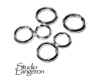 May include: A collection of silver-colored jump rings of varying sizes, scattered on a white surface. The rings are open, ready for use in jewelry making or crafts. The Studio Langeron logo is visible in the lower left corner.