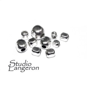 Peut inclure: Plusieurs perles brillantes de couleur argentée, de tailles variées. Les perles ont une forme géométrique arrondie avec un trou central. L'image est sur fond blanc. Le texte "studio Langeron" est visible en bas à gauche.