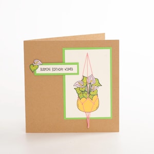 May include: A square greeting card with a kraft paper exterior. The card features a green-bordered illustration of a hanging yellow planter with white and purple flowers. The text "Blooming Birthday Wishes" is displayed on a white rectangle.