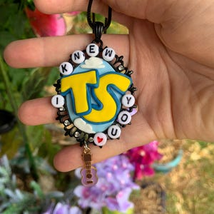May include: An oval pendant featuring a light blue background with white clouds and large yellow letters reading TS. The border consists of black filigree, small clear rhinestones, and white beads spelling I KNEW YOU with a red heart. A small guitar charm hangs below.