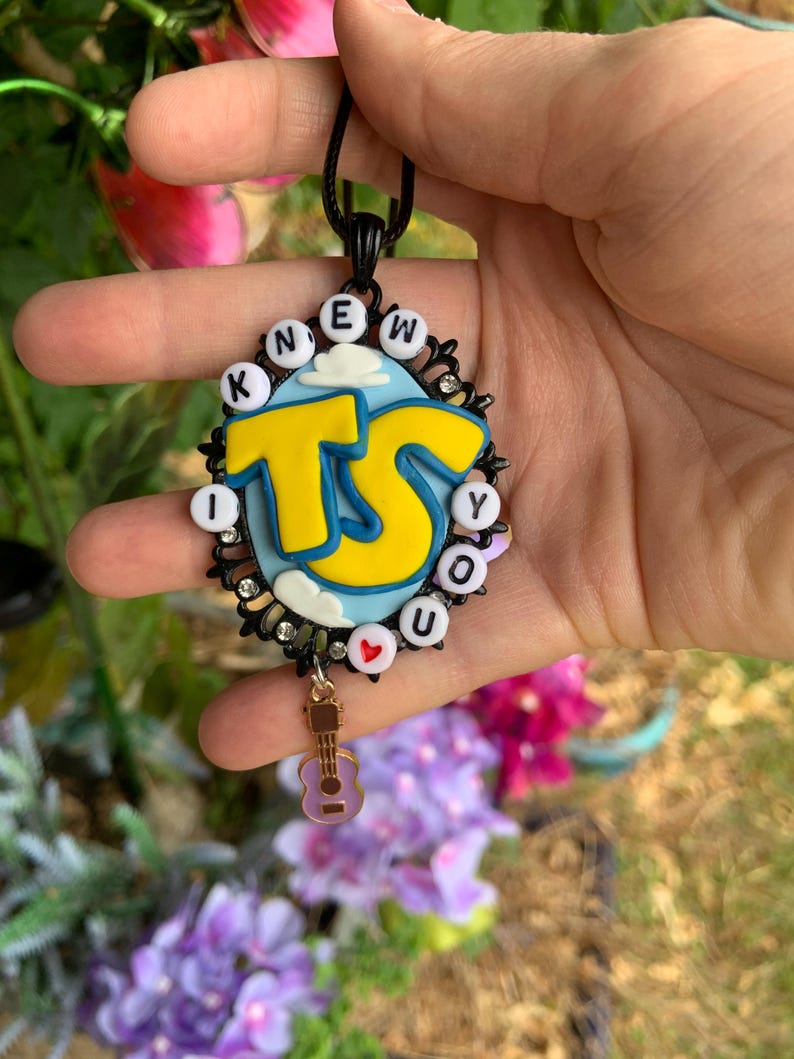 May include: An oval pendant featuring a light blue background with white clouds and large yellow letters reading TS. The border consists of black filigree, small clear rhinestones, and white beads spelling I KNEW YOU with a red heart. A small guitar charm hangs below.