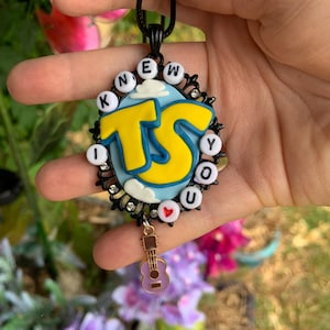 May include: An oval pendant on a black cord featuring a light blue background with white clouds and large yellow letters TS. The border is black with small clear rhinestones and white beads spelling KNEW YOU, with a small red heart and a guitar charm.