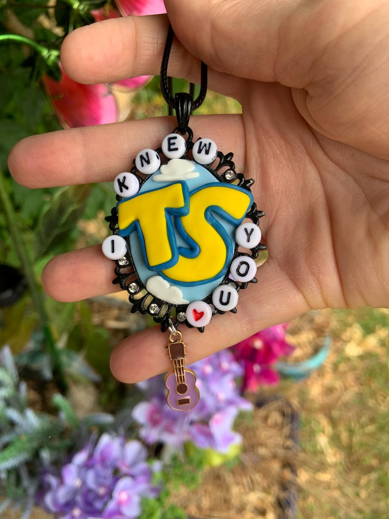 May include: An oval pendant on a black cord featuring a light blue background with white clouds and large yellow letters TS. The border is black with small clear rhinestones and white beads spelling KNEW YOU, with a small red heart and a guitar charm.