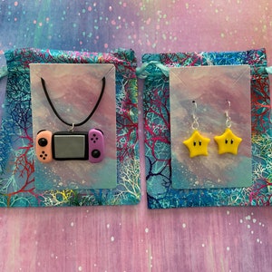 Puede incluir: Un collar con un colgante con forma de mando de videojuego y un par de pendientes con forma de estrella. El colgante es negro, rosa y melocotón. Los pendientes son amarillos con ojos negros. Las joyas se exhiben sobre un fondo azul y rosa.