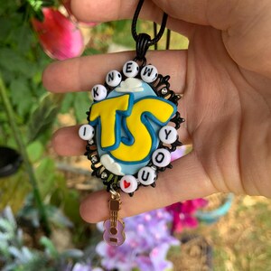 May include: A black oval pendant necklace featuring a light blue background with white clouds and large yellow letters reading TS. The border is decorated with small white letter beads spelling I KNEW YOU and a small red heart bead, plus a guitar charm.