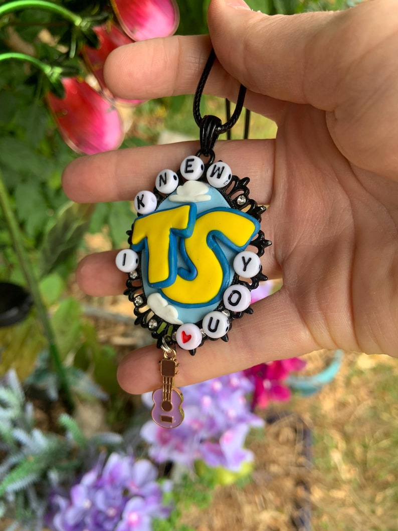 May include: A black oval pendant necklace featuring a light blue background with white clouds and large yellow letters reading TS. The border is decorated with small white letter beads spelling I KNEW YOU and a small red heart bead, plus a guitar charm.