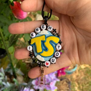 May include: An oval pendant necklace featuring a light blue background with white clouds and large yellow letters reading TS. The border is black with small clear rhinestones and white letter beads spelling I KNEW YOU. A small guitar charm hangs from the bottom.