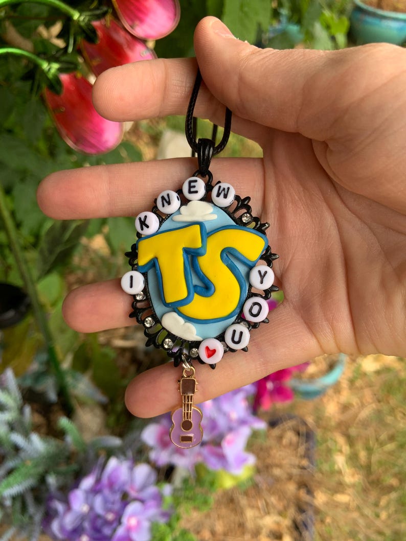 May include: An oval pendant necklace featuring a light blue background with white clouds and large yellow letters reading TS. The border is black with small clear rhinestones and white letter beads spelling I KNEW YOU. A small guitar charm hangs from the bottom.