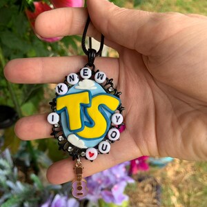 May include: An oval pendant featuring a light blue background with white clouds and large yellow letters reading TS. The border is black with small clear rhinestones and white beads spelling KNEW YOU, with a small red heart bead and a guitar charm.