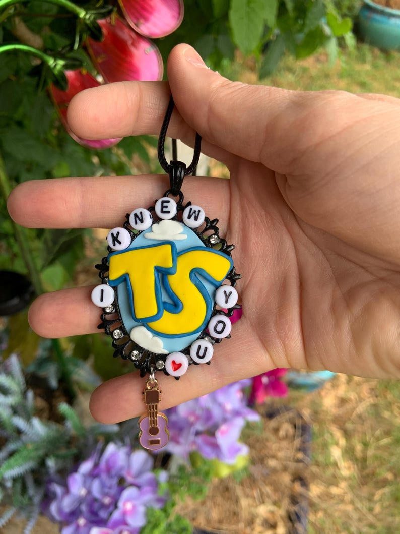 May include: An oval pendant featuring a light blue background with white clouds and large yellow letters reading TS. The border is black with small clear rhinestones and white beads spelling KNEW YOU, with a small red heart bead and a guitar charm.